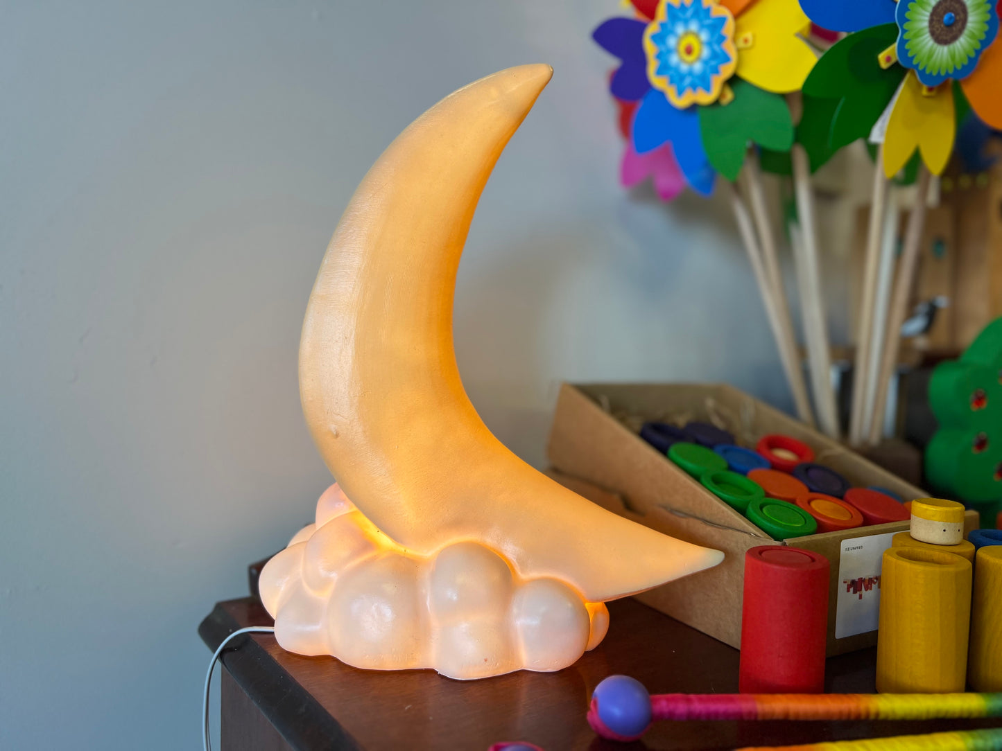 A night light in the shape of a crescent moon sits on a small cloud shape. The lightis on and gives off a warm yellow glow. There are colourful children's toys in the background.