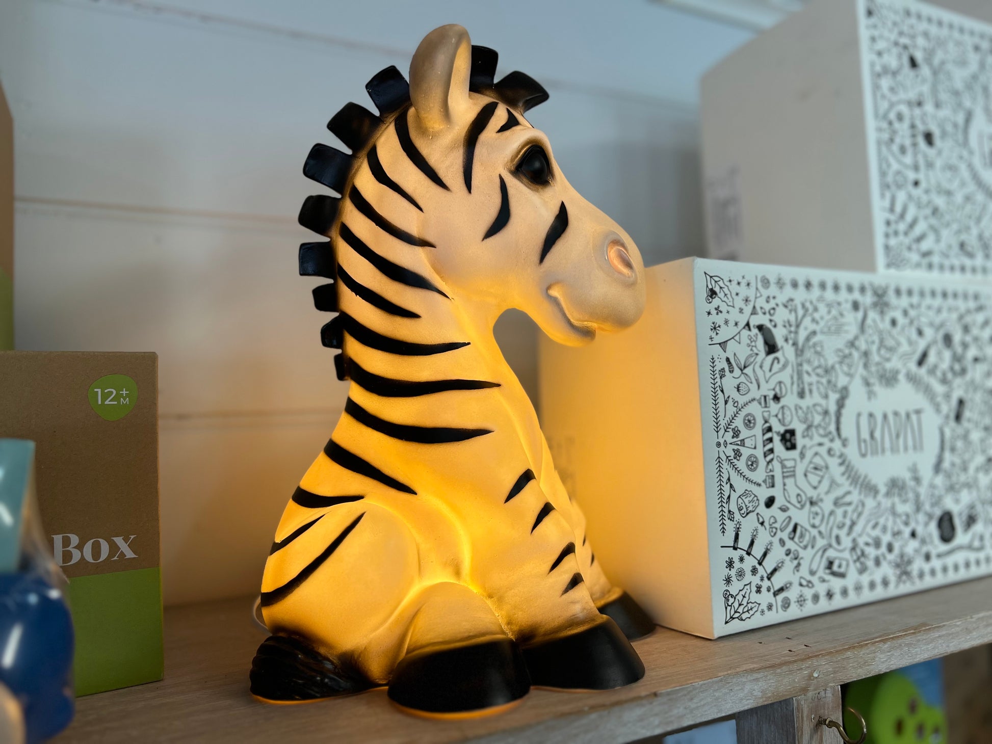 Zebra shaped nightlight facing to the right..  The light is on and shows a warm yellow glow.  The Zebra is in a sitting position.