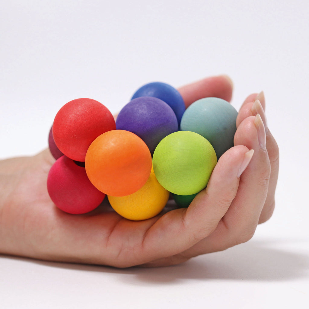 An adult hand holds a wooden bead grasper in rainbow colours