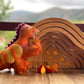 A handmade orange felt toy dragon warming itself by small wooden fire shaped pieces painted orange and yellow.  Behind the dragon is a handcrafted wooden cave made of 7 concentric pieces. The smallest piece is embellished with 7 orange sparkly gems.